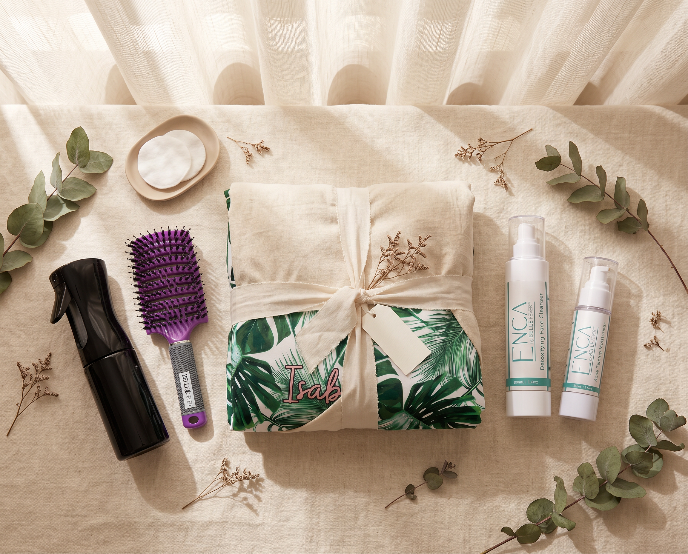 A styled flat-lay of a spa gift set on a beige linen surface with sheer curtains in the background. The set includes a cream-colored towel wrapped in a ribbon and printed with green tropical leaves and the name 'Isabel', two white ENCA pump bottles, a black spray bottle, and a purple hairbrush, surrounded by eucalyptus branches.